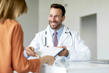 Cheerful man doctor holding clipboard and showing prescription details to female patient, discussing medical treatment with young woman