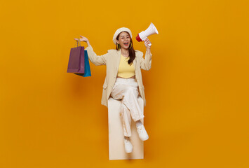 Happy Asian teen woman sitting on chair holding shopping bags and announcement discount in megaphone on yellow studio background. happy shopping summer sale.