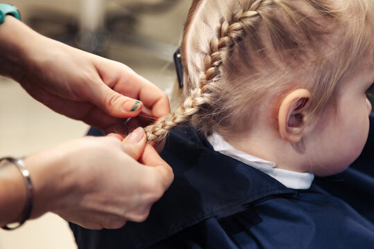Closeup of hairstylist braiding braids for kid at barbershop at mirror. Hair salon, barber woman make hairstyle for cute little blonde girl in barber shop. Haircare, beauty concept. Copy ad text space