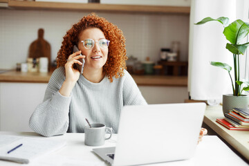 Female accountant in casual clothes and glasses working from home on laptop, making phone call to bank concerning information of financial account of company, sitting at kitchen table with laptop - 619194733