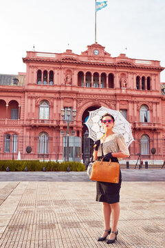 caucasian actrees woman dressed in vintage clothes and umbrella in the street with The Casa Rosada in the background	