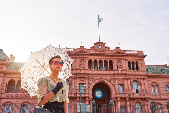 caucasian actrees woman dressed in vintage clothes and umbrella in the street with The Casa Rosada in the background