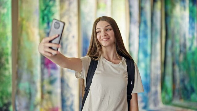Young beautiful girl student wearing backpack make selfie by smartphone at art gallery