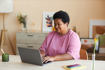 Happy mature African American woman in pink t-shirt sitting by desk in front of laptop in living room, typing on keyboard and networking