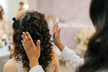 Close-up rear view of unrecognizable hairdresser applying hair styling product to female client curly hairstyle in beauty studio. Concept of professional fashion hairdresser and hair beauty salon.