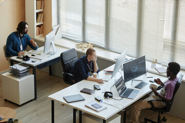 Young intercultural coworkers having snacks while sitting by their workplaces in front of computer screens at lunch break in office