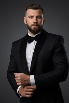 Confident and stylish handsome man with a manicured beard wears a luxurious black suit and bowtie on a grey background