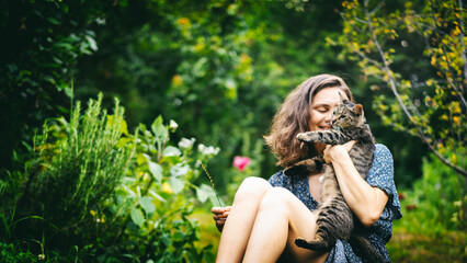 Young cheerful smiling woman holding in her arms a cute gray cat sitting on the grass in a summer garden