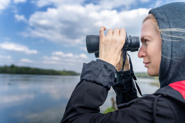 Caucasian middle aged woman traveler looking through binoculars while walking on the river bank