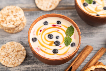 Yoghurt with bilberry and caramel in clay bowl on gray wooden, side view, selective focus.