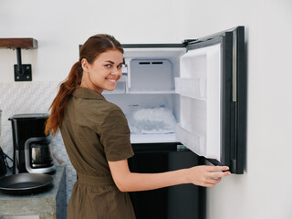 Woman smiling with teeth looking into camera in kitchen at home opened freezer empty with ice inside, home refrigerator, defrosted, view from back.