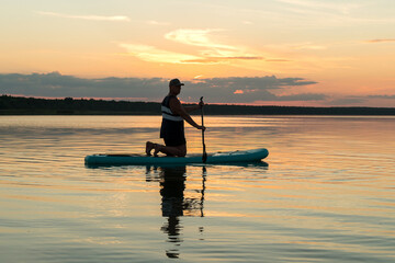 A man in shorts on his knees on a sapboard with an oar at sunset swims in the water of the lake against the backdrop of a pink sky reflection in the water.