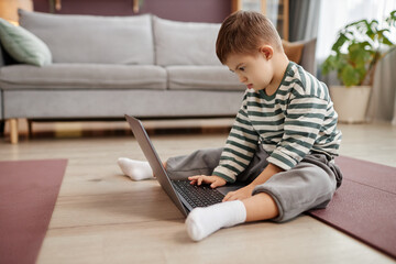 Full length side view portrait of little boy with down syndrome using laptop on floor at home, copy...