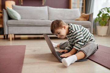 Naklejka premium Full length side view portrait of curious little boy with down syndrome using laptop while sitting on floor at home, copy space