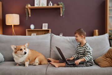 Side view portrait of curious little boy with down syndrome using laptop computer while sitting on comfortable sofa with dog, copy space