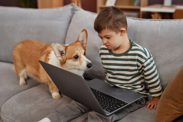 Portrait of little boy with down syndrome using laptop while sitting on sofa with cute dog watching