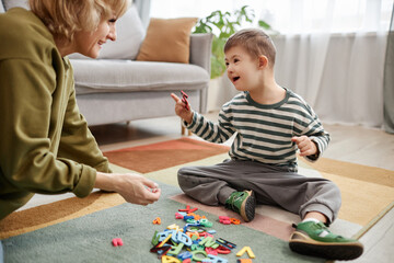 Naklejka premium Full length portrait of happy little boy with down syndrome playing on floor and enjoying learning exercises with mother