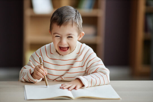 Portrait of excited little boy with down syndrome drawing pictures while sitting at table and laughing happily, copy space