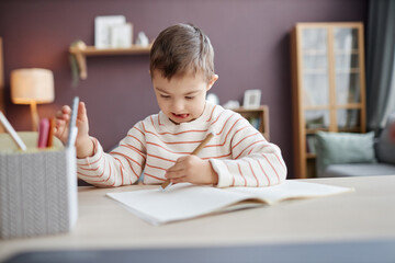 Front view portrait of focused little boy with down syndrome drawing pictures sitting at table at home