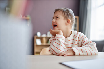 Side view portrait of happy little boy with down syndrome sitting at desk in school and laughing,...
