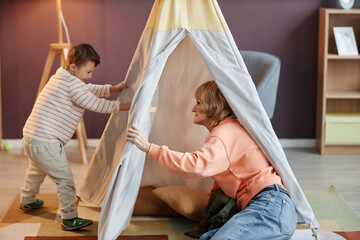 Full length portrait of little boy with down syndrome playing with mother in play tent at home
