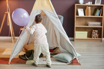 Back view of cute little boy playing with tent at home and holding colorful balloon, copy space