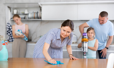 Smiling tidy young housewife cleaning apartment together with her husband and daughters, assiduously wiping kitchen table