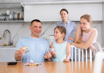 Cheerful adult man sitting at table in kitchen and counting money while wife doing household chores and two teenage daughters standing nearby waiting for pocket money..