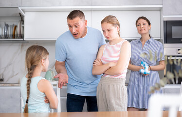 Obraz premium Displeased mother and father reprimanding preteen girl while standing with older sister in home kitchen. Conflict between parents and children