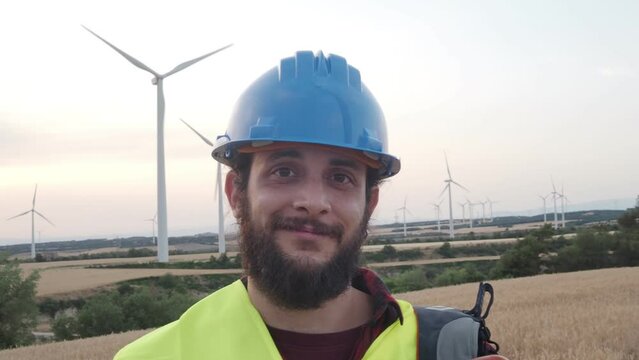 Worker man with helmet looking at camera smiling in a field of windmills