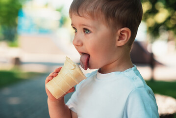 A cute little European-looking kid is eating an ice cream cone in a waffle cup in the park. Close-up. Summer time, happy