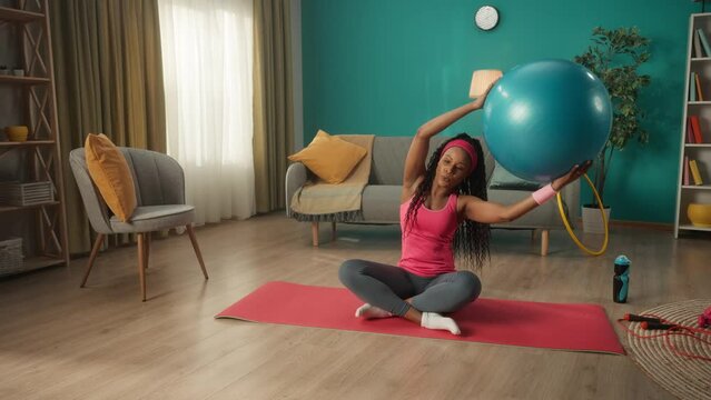 A young woman is sitting cross legged on a sports mat, doing a press and arm exercise. African American woman raises straight arms with a fitness ball above her head and does side bends.