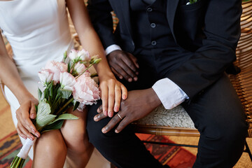 Closeup of African American bride and groom sitting on bench together and holding hands with focus on wedding rings, copy space - Powered by Adobe