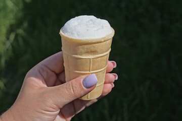one white ice cream in a brown waffle cup is held by a girls hand on the street on a green...