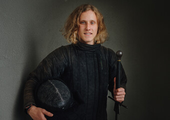 Portrait of a smiling long-haired young teen dressed in black historical fencing armor with a long medieval sharp sword