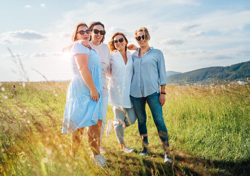 Portrait of four cheerful smiling and laughing women embracing during outdoor walking by high green grass hill. They looking at the camera. Woman friendship, relations, and happiness concept image.