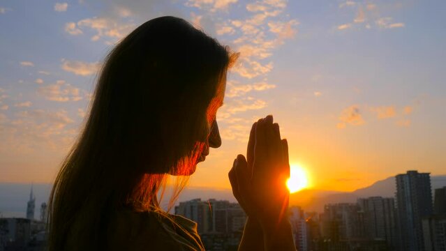 Young woman silhouette is folding hands and praying against the sunrise or sunset sky over the city - close up side view, sun lens flares. Religion, prayer gesture, hope, spiritual and urban concept