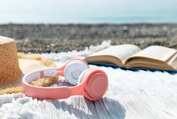 Audiobook or podcast during summer holidays. Close up of headphones near open book, sunglasses and hat on the beach with seaside in background.