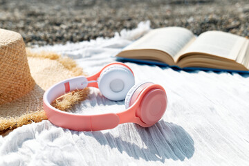 Audiobook or podcast during summer holidays. Close up of headphones near open book, sunglasses and hat on the beach with seaside in background.
