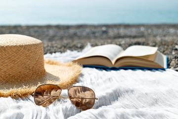 Open book, sunglasses and straw hat on the towel on pebble beach. Concept of reading and relaxing in summer vacation. Beach literature.