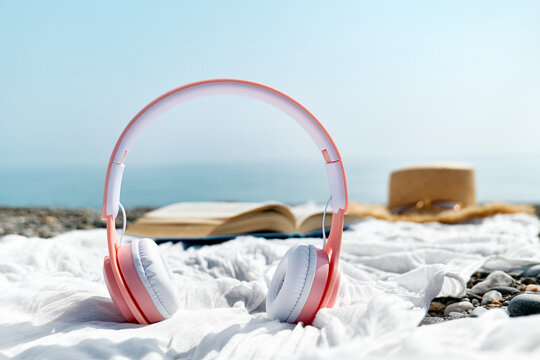 Audiobook or podcast during summer holidays. Close up of headphones near open book, sunglasses and hat on the beach with seaside in background.