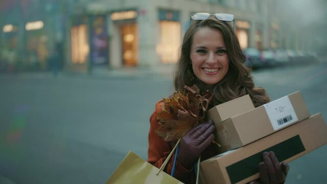Hello september. smiling trendy middle aged woman in orange trench coat with parcels, shopping bags and autumn yellow leaves near shop in the city.