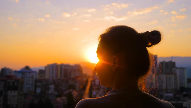 Back view: woman is standing on the balcony and looking at the sunset, sunrise sky over the city - close up, sun lens flares. Lonely, urban, dramatic and freedom concept