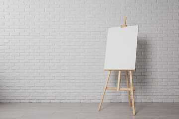 Wooden easel with blank canvas near white brick wall indoors. Space for text