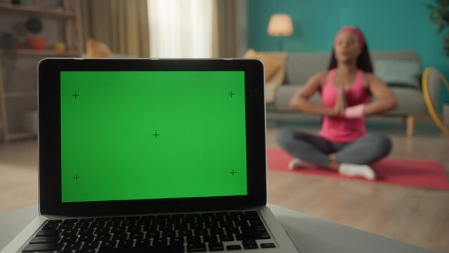 A laptop with a green screen on a table against a blurred background in a room close up. In the background, a woman sits in a lotus position on the floor. Advertising area, workspace mock up.