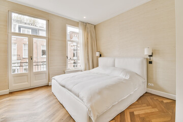 a bedroom with wood flooring and white bed in front of two windows looking out onto an apartment balcony area