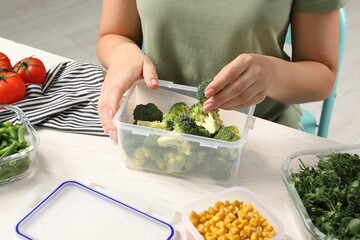Woman putting broccoli into plastic container at white wooden table, closeup. Food storage