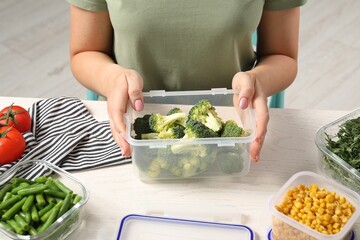 Woman holding plastic container with broccoli at white wooden table, closeup. Food storage