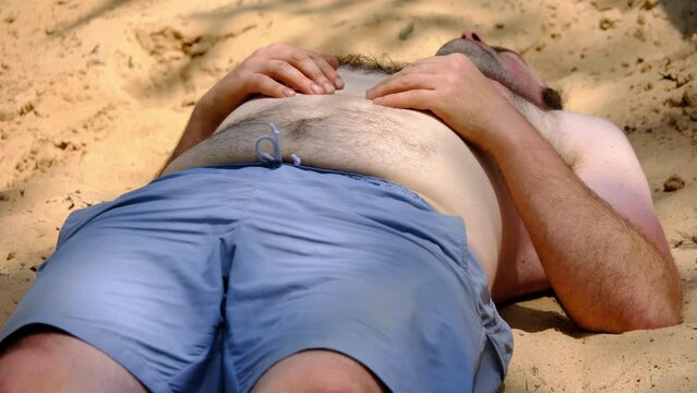 Close up of slightly obese man with blue shorts and topless with hairy chest waking up and stretching after falling asleep on top of pile of sand after back yard pool party.