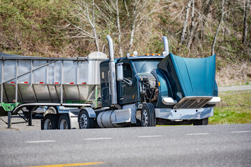 Dark green broken big rig day cab semi truck with open hood and bulk semi trailer standing out of service on the road shoulder waiting for mobile towing truck